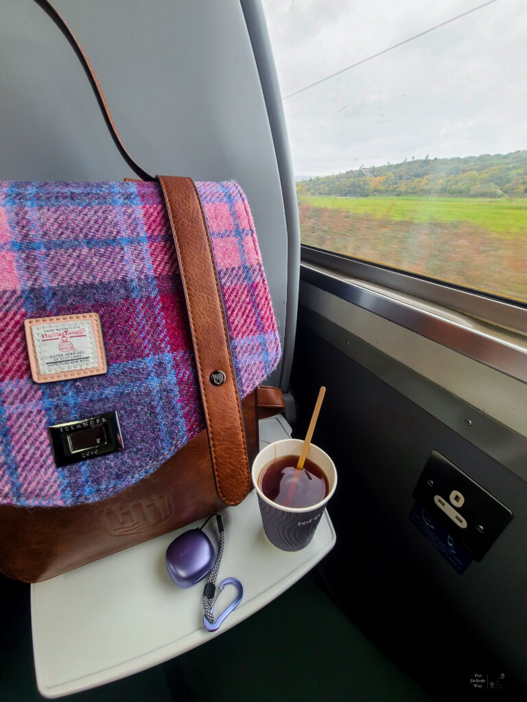 Backpack, tea, and earbuds on train tray table with the few of the Scottish Highlands outside the window - taking the train is a great way to help curb travel anxiety for me